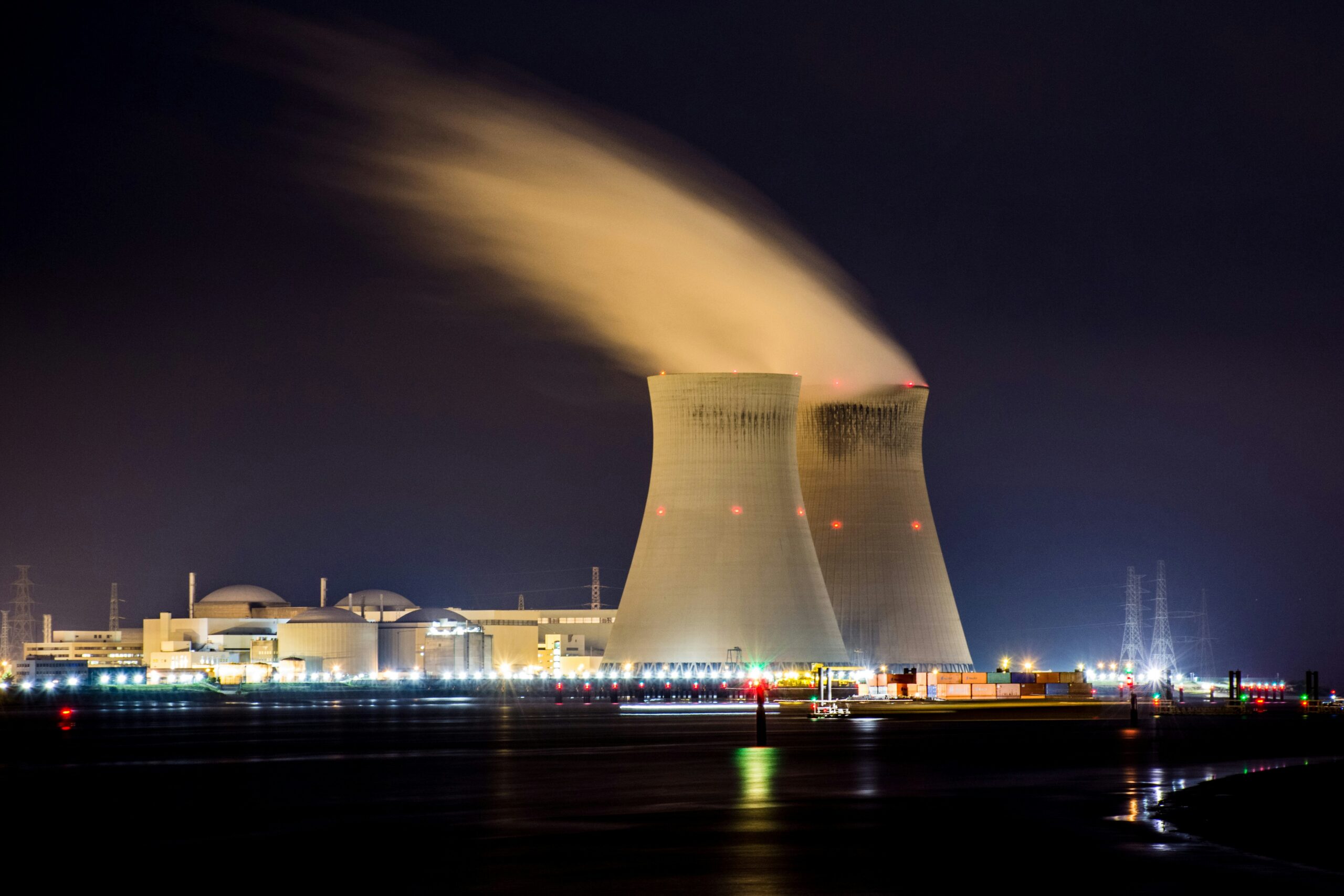 Nuclear power plant at night with two illuminated cooling towers releasing steam plumes, reactor containment domes and high-voltage transmission lines visible in the background.